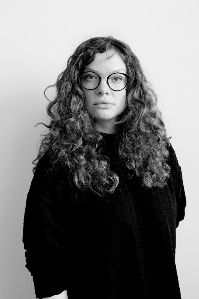 Young woman with long curled hair and glasses stands tall against a white wall.
