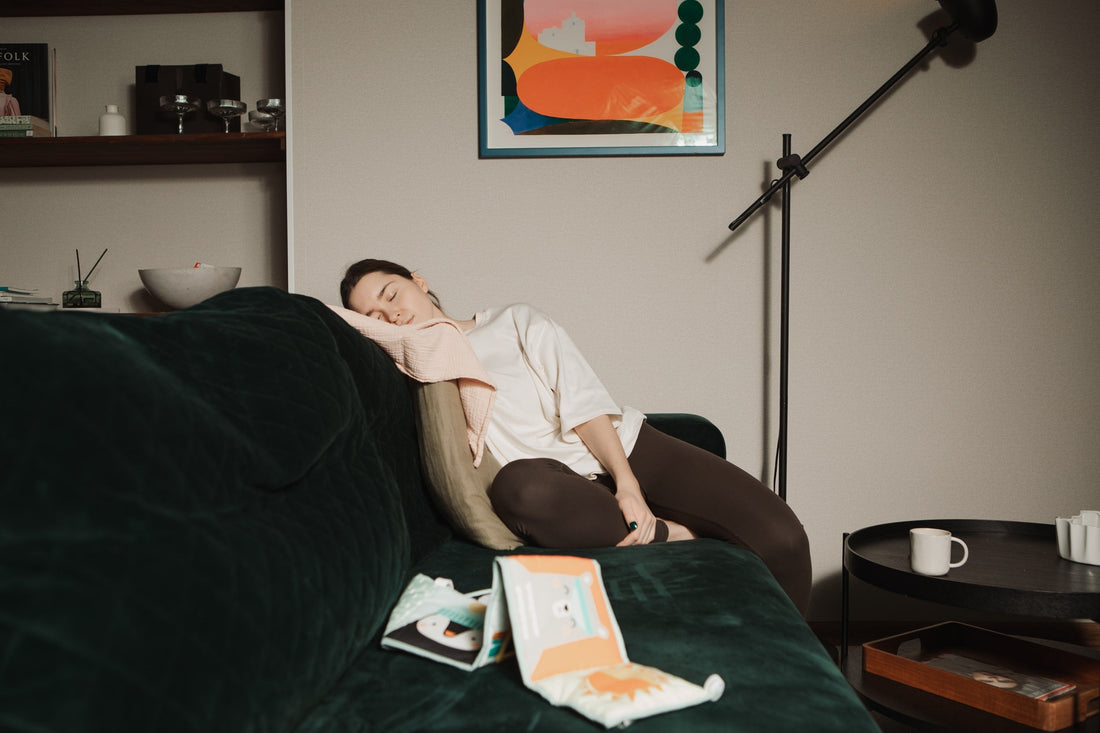 A woman is sleeping on a dark green velvet couch in a cozy living room. She is wearing a loose white top and dark leggings, resting her head on a pillow with a pink cloth draped over it. Children's books or baby items are scattered on the couch near her. 