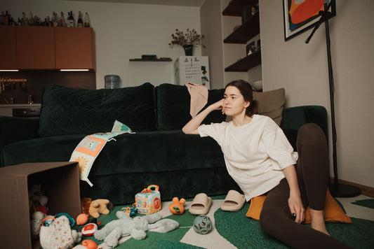 A young mother wearing a white oversized nursing t-shirt and dark leggings sits on the floor in front of a green couch, resting her head on one arm. The living room floor around her is scattered with colorful baby toys, and she looks tired.