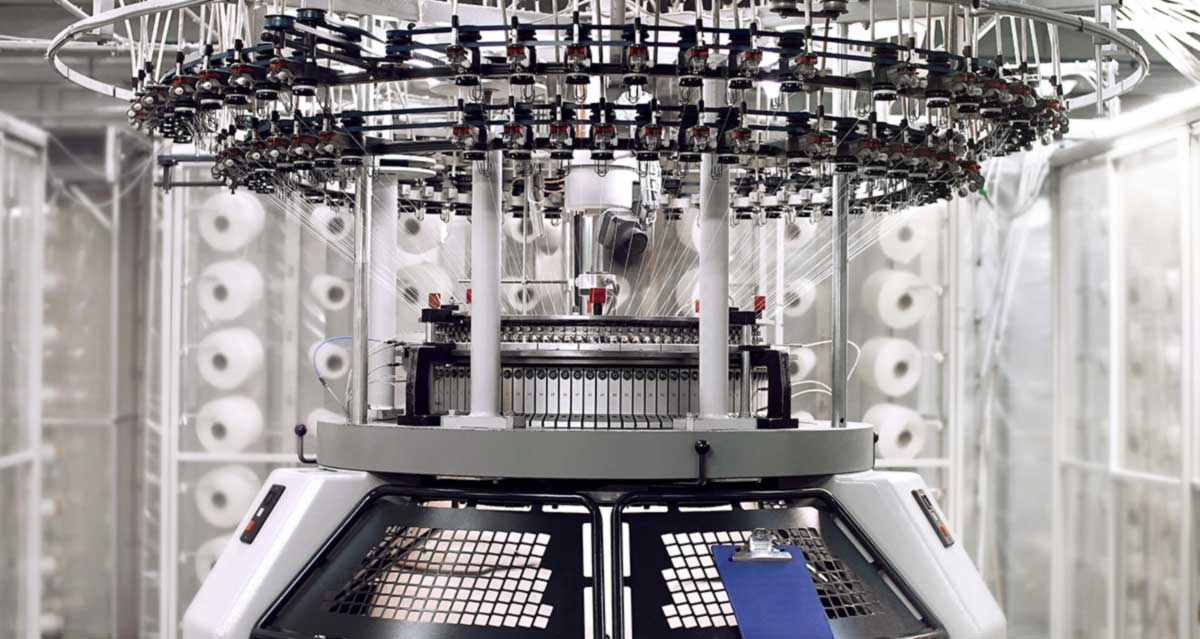 A close-up of a circular knitting machine creating white organic cotton fabric.