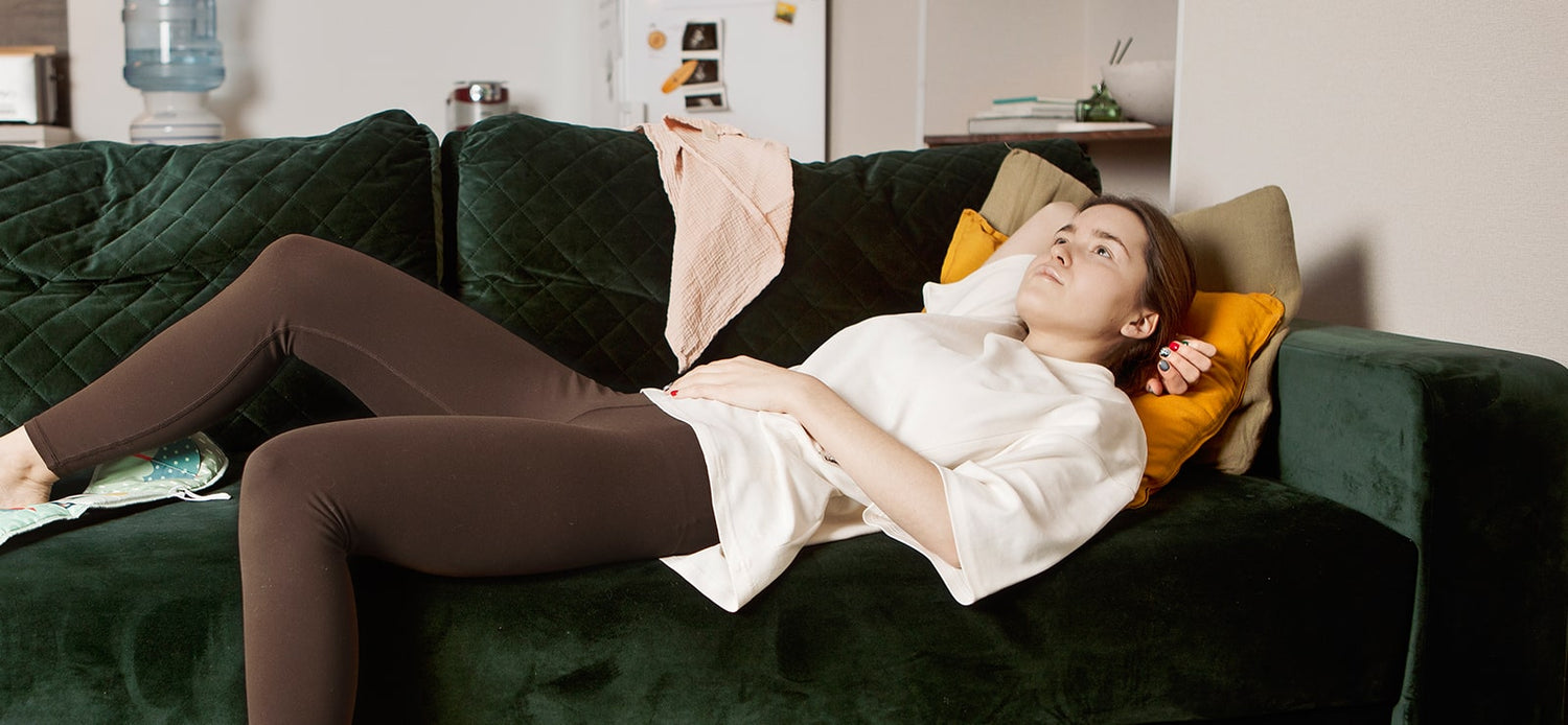 Mother relaxing on a couch in a white nursing t-shirt and leggings, surrounded by baby toys.