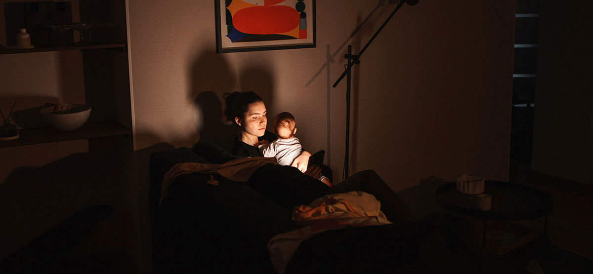 Young mother sitting on a sofa with her baby cradled on her chest, scrolling through her smartphone in a dimly lit living room.