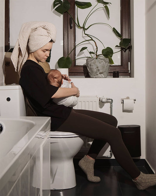 Mother sitting on the toilet holding her newborn, wearing a black nursing t-shirt, reflecting real motherhood.