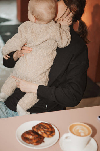 Mother wearing a black nursing jumper, sitting at a café table with her baby standing on her lap, holding him lovingly. Comfortable and stylish breastfeeding clothes for nursing moms.