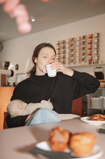 Mother wearing a black nursing jumper, sitting in a café, sipping coffee while discreetly breastfeeding her baby. Stylish, functional nursing clothes and breastfeeding top for on-the-go moms.