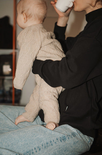 Mother wearing a black nursing jumper, sitting in a café, sipping coffee while holding her baby standing on her lap. Stylish and practical breastfeeding clothes for nursing moms on the go.