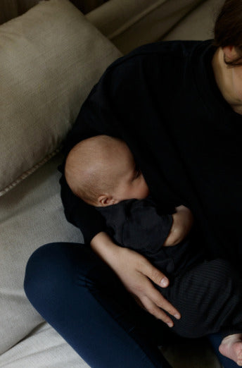 Mother sitting on a couch wearing a black nursing jumper, breastfeeding her newborn. Cozy and functional nursing clothes for breastfeeding moms.