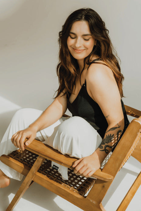 Happy woman sitting in a lounge chair wearing a black nursing top and white pants. Modern, stylish, and comfortable breastfeeding clothes for nursing moms.