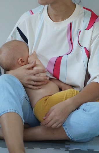 Mother sitting on a playmat breastfeeding her baby while wearing a colorful nursing t-shirt. Comfortable and stylish breastfeeding clothes perfect for nursing moms.