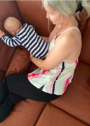 Mother wearing a colorful nursing top playing with her child on a brown leather couch. Comfortable and stylish breastfeeding clothes for nursing moms.

