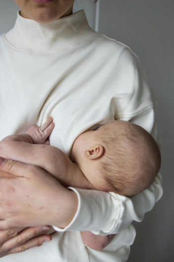 Mother holding her baby in her lap, wearing a modern white nursing jumper. Stylish and comfortable breastfeeding clothes for nursing moms.