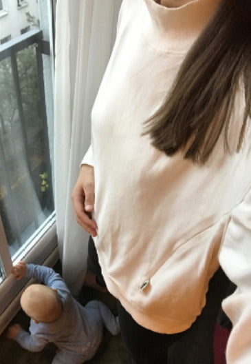 Young mother wearing a modern, cool white nursing jumper, standing by the window and caring for her little one. Stylish and comfortable breastfeeding clothes perfect for nursing moms.