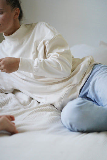 Young mother wearing jeans and a white nursing jumper, sitting on a bed and lovingly caring for her little one. Comfortable and stylish nursing clothes ideal for breastfeeding moms.