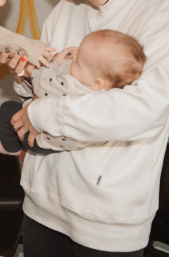 Close-up of a standing mother wearing a white nursing jumper, holding her baby while another baby reaches out to touch him. Comfortable and stylish nursing clothes for breastfeeding moms.