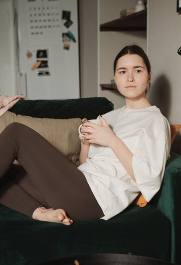 Young woman sitting on a couch holding a tea cup, wearing a white nursing t-shirt. Stylish and comfortable breastfeeding clothes for nursing moms.
