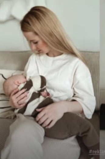 Young mother wearing an oversized white nursing tee, breastfeeding her baby on her lap while sitting on a couch. Comfortable and stylish nursing clothes for breastfeeding moms.