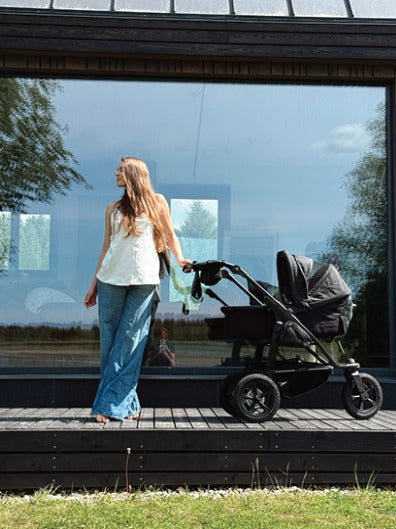 Mother standing on her porch wearing a white organic cotton nursing top and blue jeans, holding a baby stroller. Stylish and eco-friendly breastfeeding clothes for modern nursing moms.