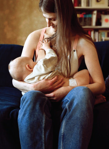 Mother wearing a white nursing top, sitting on a couch and breastfeeding her child. Comfortable and stylish breastfeeding clothes for nursing moms.